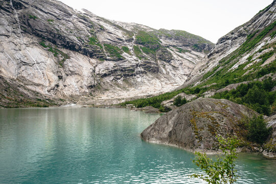 Turquoise glacial lake at the base of grey granite mountains with waterfalls cascading down cliffs under a cloudy sky in a wilderness area.