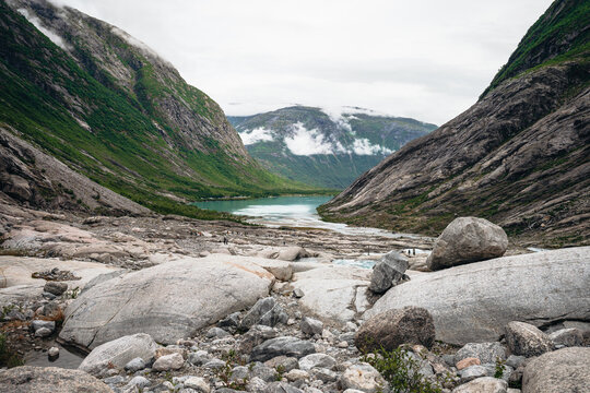 A wide-angle view of a rocky glacial valley leading to a turquoise lake in Norway. Hikers are visible on a path and bridge under a cloudy sky between mountains.
