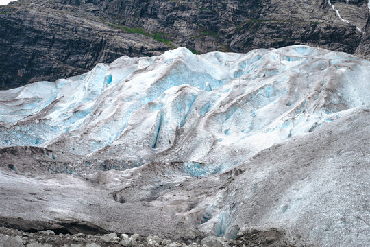 Detailed view of a massive blue glacier showing deep crevasses and textured ice formations against a dark rocky mountain wall in a cold outdoor environment.