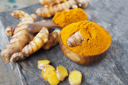 Fresh turmeric roots with sliced pieces and turmeric powder in wooden bowl on rustic wooden background.