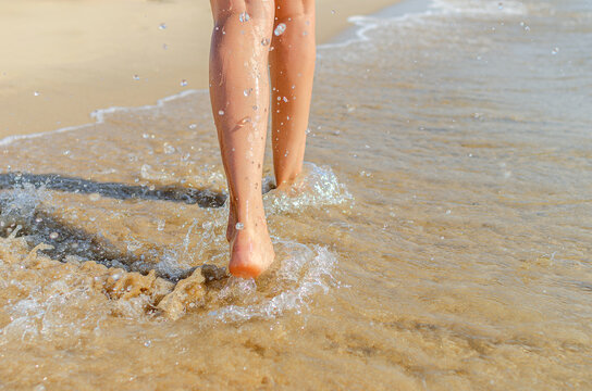A woman walks barefoot along the sandy ocean beach as waves splash water around her feet.
