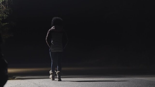 Woman walks on a dark road. The person is wearing a jacket and has a backpack. The scene is dark and moody, with the person standing alone in the middle of the road