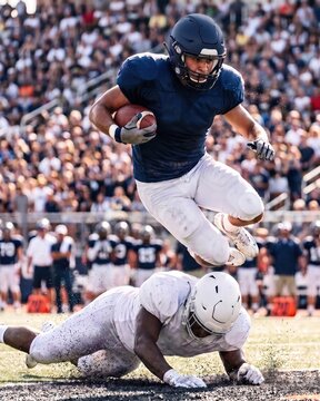 Fearless ball carrier leaps over diving tackler. Concept of agility, obstacle avoidance, bold action, athletic skill, and maintaining forward progress.