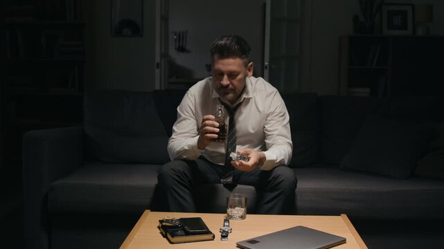 Middle aged Caucasian businessman pouring whiskey from bottle into glass while suffering from stress. Perfect for commercial projects on alcohol abuse, mental wellbeing and corporate burnout