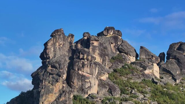 Rugged mountain range with jagged, weathered rock formations and sparse green vegetation under a clear blue sky. Gradual zoom reveals intricate erosion patterns and subtle cloud shadows drifting
