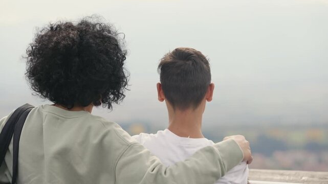 Two people are standing on a balcony looking at panoramic view over a city. One of them is holding a cell phone