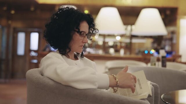 A woman is sitting in a chair reading a book. She is wearing glasses and has a book in her hand. The scene is set in a restaurant with a few other chairs and tables around her