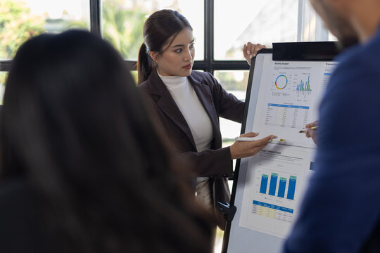 Businesswoman presenting financial data at office meeting. Business team collaborating during a presentation, analyzing charts and graphs on a whiteboard and table in an office.