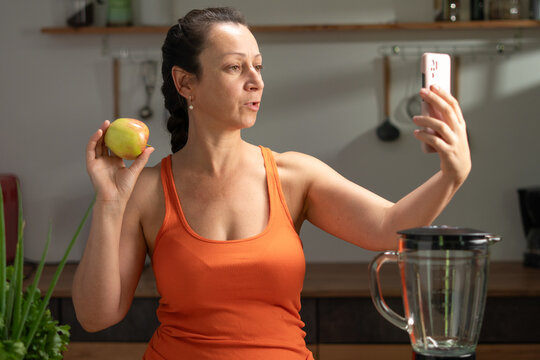 Young woman eating apple as healthy food and taking selfie in kitchen