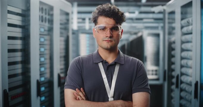 Young Male IT Specialist in Safety Glasses Stands with Arms Crossed inside Modern Data Center Server Room. System Administration and Cloud Storage Infrastructure Ensure Data Security. Portrait.