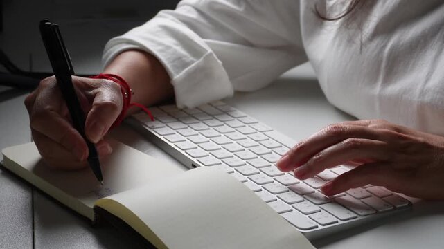 Businesswoman's hands taking handwritten notes in a journal while simultaneously typing on a computer keyboard, concept of multitasking