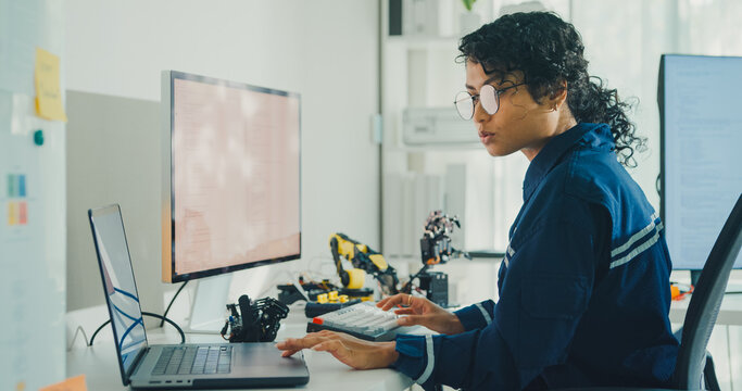 Young African American female robotics engineer coding AI software for robotic arm in laboratory. Embedded system, firmware testing, machine learning, industrial automation, future technology concept.