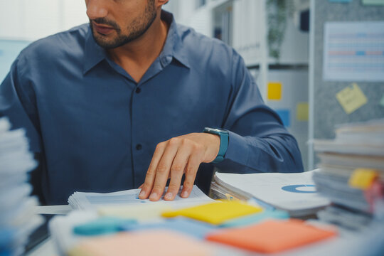 Closeup overworked young Latin businessman reading document in large stack of paperwork file at office desk. Tax audit deadline, accounting stress, corporate burnout, bookkeeping, bureaucracy concept.