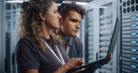 Obraz premium Focused Female System Administrator Types on Laptop While Colleague Conducts Server Cabinet Check in Data Center Corridor. Women in STEM Drive Modern Enterprise IT Operations and Cloud Security.
