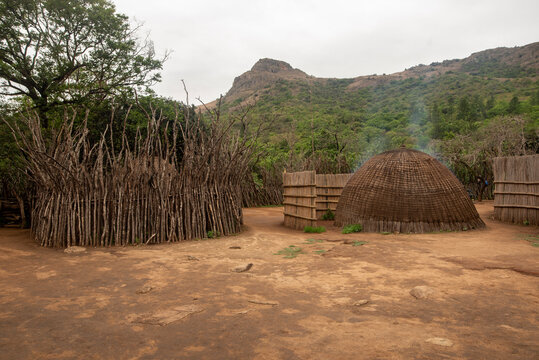 A wide shot displays a traditional Swazi homestead featuring thatched beehive huts, woven reed fences, and a wooden cattle kraal set against a backdrop of green mountains in Eswatini.