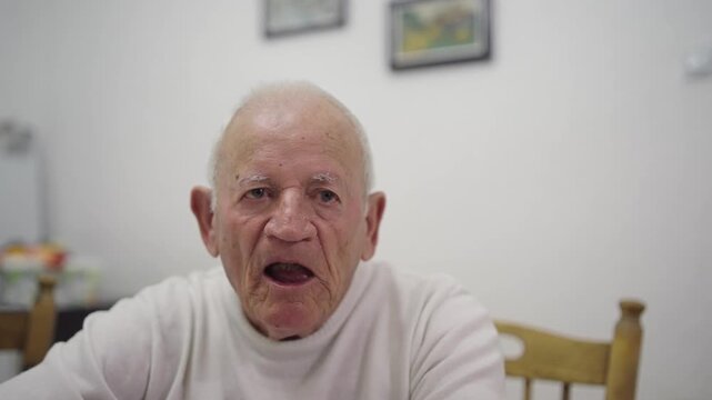 Thoughtful old man with a white shirt and a beard is sitting at a table looking at  the camera with a serious expression