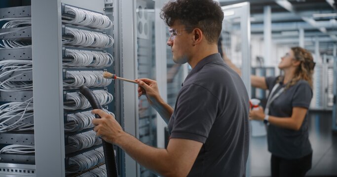 In Data Center, Male Technician Vacuums Dust From Server Cables With Brush While Female Colleague Cleans Cabinet Glass. Professional IT Facility Maintenance Guarantees Enterprise Server Reliability.