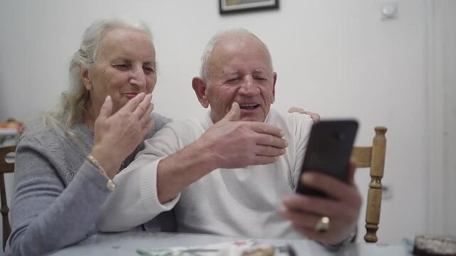 Happy smiling old man and woman couple are sitting together and looking at a cell phone while making an online call