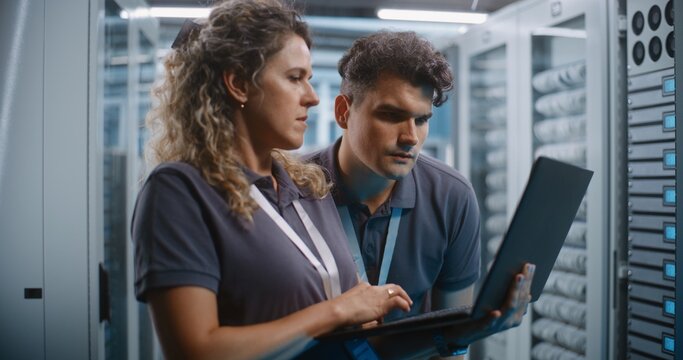 Female IT Specialist Operates Laptop While Teammate Examines Server Rack Hardware Modules in Background. Combined Physical and Software Maintenance Ensures Seamless Cloud Infrastructure Operations.