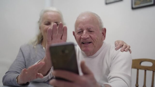 Happy smiling old man and woman couple are sitting together and looking at a cell phone while making an online call