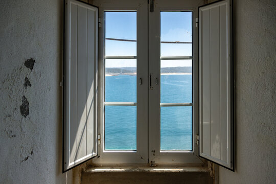 Sea view through an open window at Sao Miguel Arcanjo Fort in Nazare, Portugal, revealing the blue Atlantic and distant coastline. A historic interior framing a scenic coastal landscape.