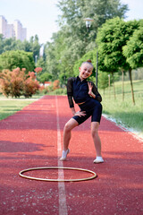 A young girl wearing gym attire poses cheerfully on a running track with a hoop nearby in a vibrant park scene. The background features lush greenery, colorful plants, and distant buildings. © Loginova