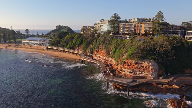 Terrigal coastal boardwalk winds beside sandstone cliffs and ocean at sunrise, drone backdrop as people walk and enjoy the day as waves crash on shore