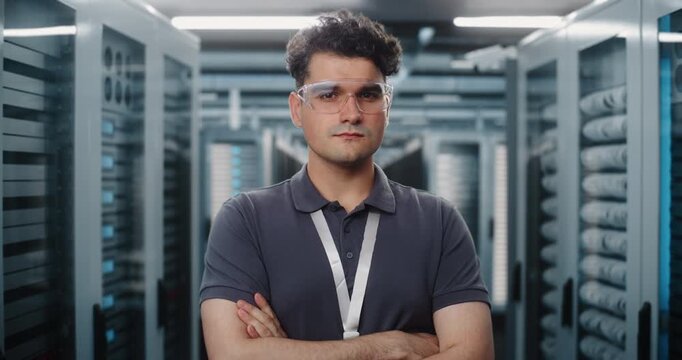 In Modern High Tech Data Center, Male Network Engineer Poses Confidently Between Server Racks. Reliable Hardware Infrastructure and Cybersecurity Systems Protect Enterprise Digital Assets. Portrait.