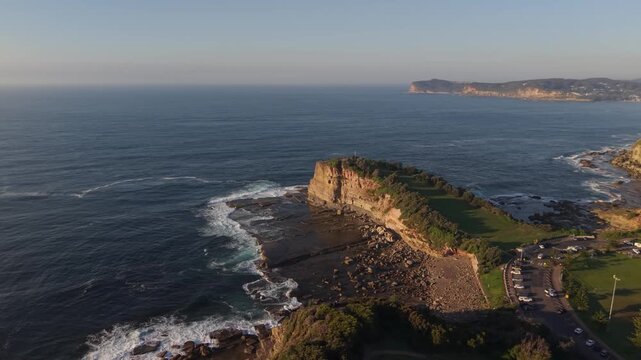Terrigal Skillion headland rises above rocky ocean shoreline at sunrise, drone medium orbit backdrop