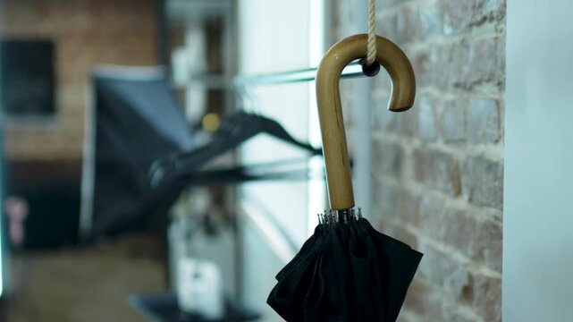 Curved wooden umbrella handle hanging from hook in indoor entryway on light brick wall with folded black umbrella canopy under soft daylight close up still life composition
