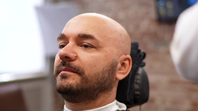 Relaxed bald man in barber chair as barber moves electric clippers along sideburn and jawline, close facial grooming with steady hands and calm expression