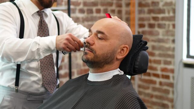 Barber trimming beard with electric clipper on reclined bald man in brick barbershop with soft natural side light