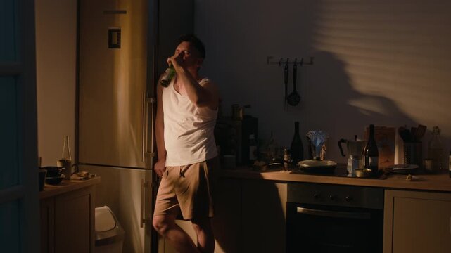 Middle aged Caucasian man drinking alcohol from bottle alone in kitchen. Useful for addiction recovery campaigns, mental health awareness and social issue videos