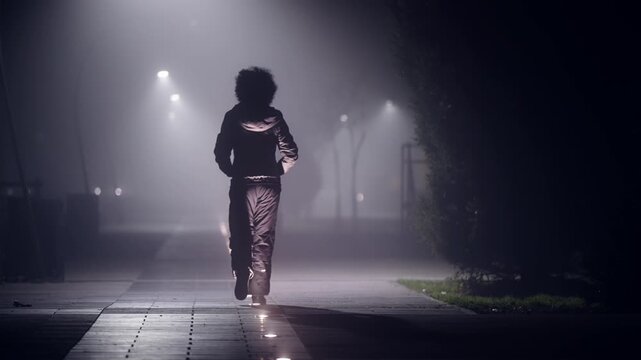 A woman is walking down a sidewalk pavement in the dark foggy time. The street is lit by street lamps. The woman is wearing a white jacket and she is alone. The scene is quiet and peaceful