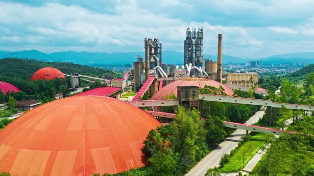Aerial view of a cement plant with massive orange domes, metal processing towers, elevated conveyors, forested hills, and distant mountains beneath blue clouds in soft light.