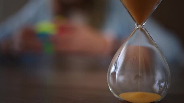 Conceptual video of a classic sandglass measuring the passage of time on a wooden table, with an out-of-focus person in the background solving a colorful puzzle cube as a metaphor for deadlines