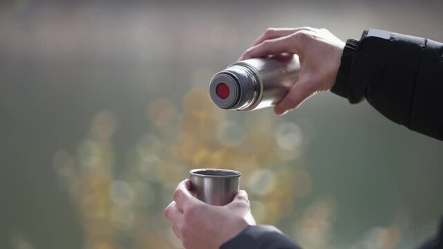 A person is pouring hot water into a thermos. The person is wearing a black jacket and he is outdoors