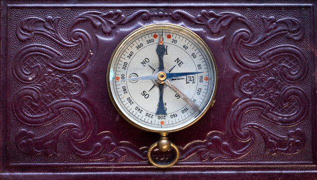 Antique old compass resting on a richly decorated embossed book, featuring intricate baroque and gothic ornamentation, aged metal details, dark luxurious tones, and a historical vintage atmosphere.
