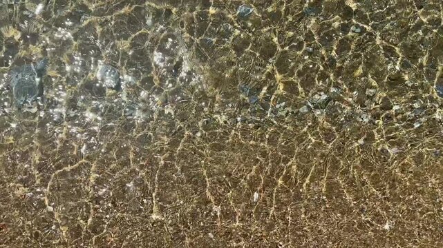 Top-down close-up of clear shallow water rippling over pebbles and small rocks. Sunlight creates shimmering patterns across the submerged stones and patches of green algae.