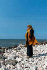 Woman wearing yellow hijab and sunglasses stands posing on white rocky beach coast day. © wahyu