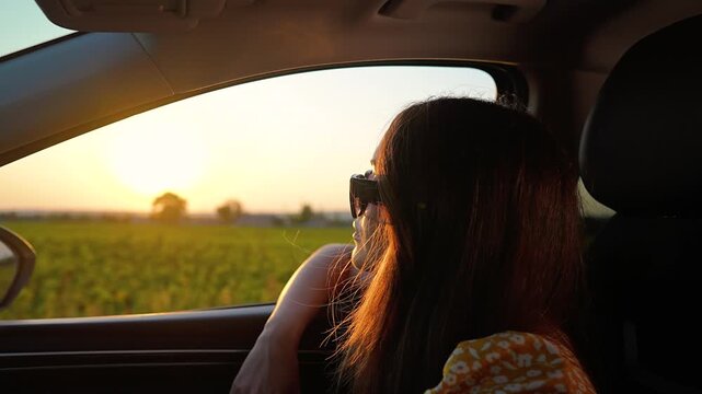 Young woman looks out car window during sunset travel. Girl enjoys summer road trip in moving car. Female passenger watches sunset view through window. Happy woman loves travel vacation drive.