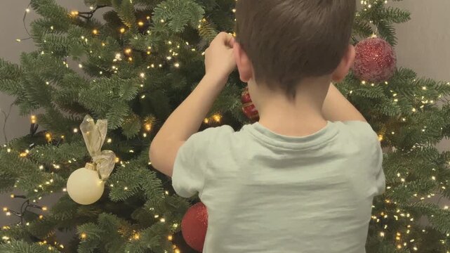 Boy placing ornament on Christmas tree among lights. Classic Christmas family moment. 