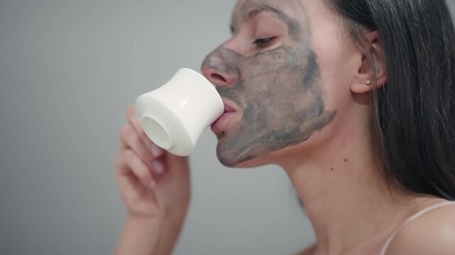 A woman with face mask is holding a cup in her hand drinking coffee. Concept of relaxation and self-care, as the woman takes the time to pamper herself with a face mask