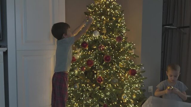 Child stretches to reach top of Christmas tree while younger sibling helps with decorations. Family teamwork and holiday preparation concept.