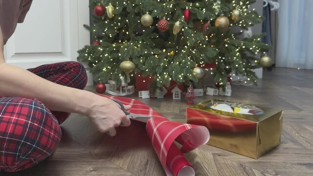 Hands cutting wrapping paper on floor near Christmas tree preparing gifts. Cozy home atmosphere and festive preparation. Suitable for holiday marketing, ecommerce and seasonal content. 