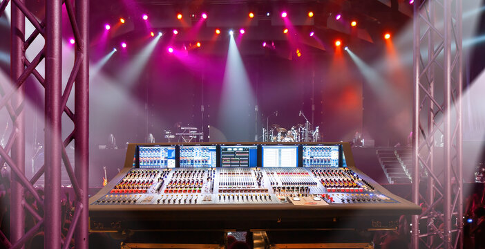 Concert production, professional audio mixing console controls live sound while colorful stage lighting illuminates a music performance venue.
