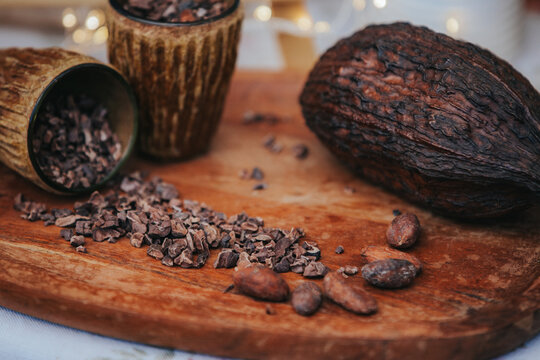 Cacao pod and cocoa nibs displayed on wooden cutting board with rustic cups, showcasing natural ingredients for chocolate production in a warm, inviting setting