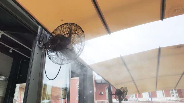 Outdoor misting fan sprays fine water mist under terrace awning. Commercial misting fan cools outdoor cafe patio with fine water vapour stream on hot summer day for comfortable climate control.