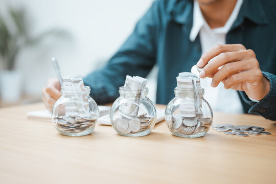 Savings grow through a deposit and a plan for financial allocation as a hand adds a coin to one jar of three, dividing savings by each deposit under a plan for financial allocation beside a notebook.
