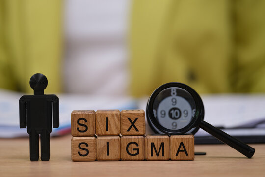 A desk setup features wooden blocks spelling SIX SIGMA, alongside a magnifying glass and a small figure.